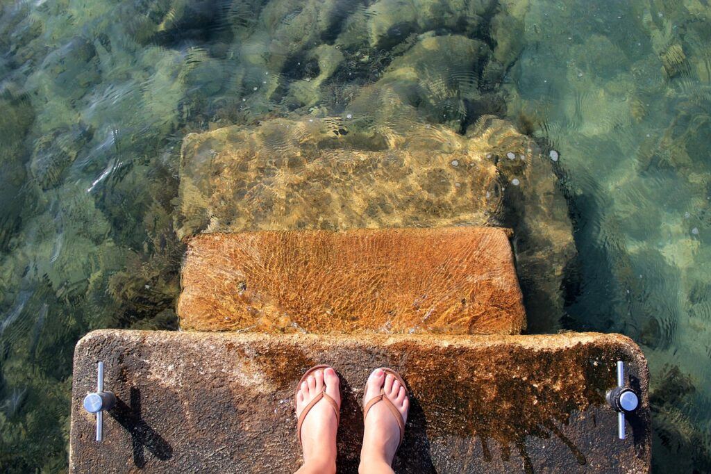 Taking action steps by taking the plunge. a pair of feet, standing on stone steps, looking down at the sea water that is a few steps below.